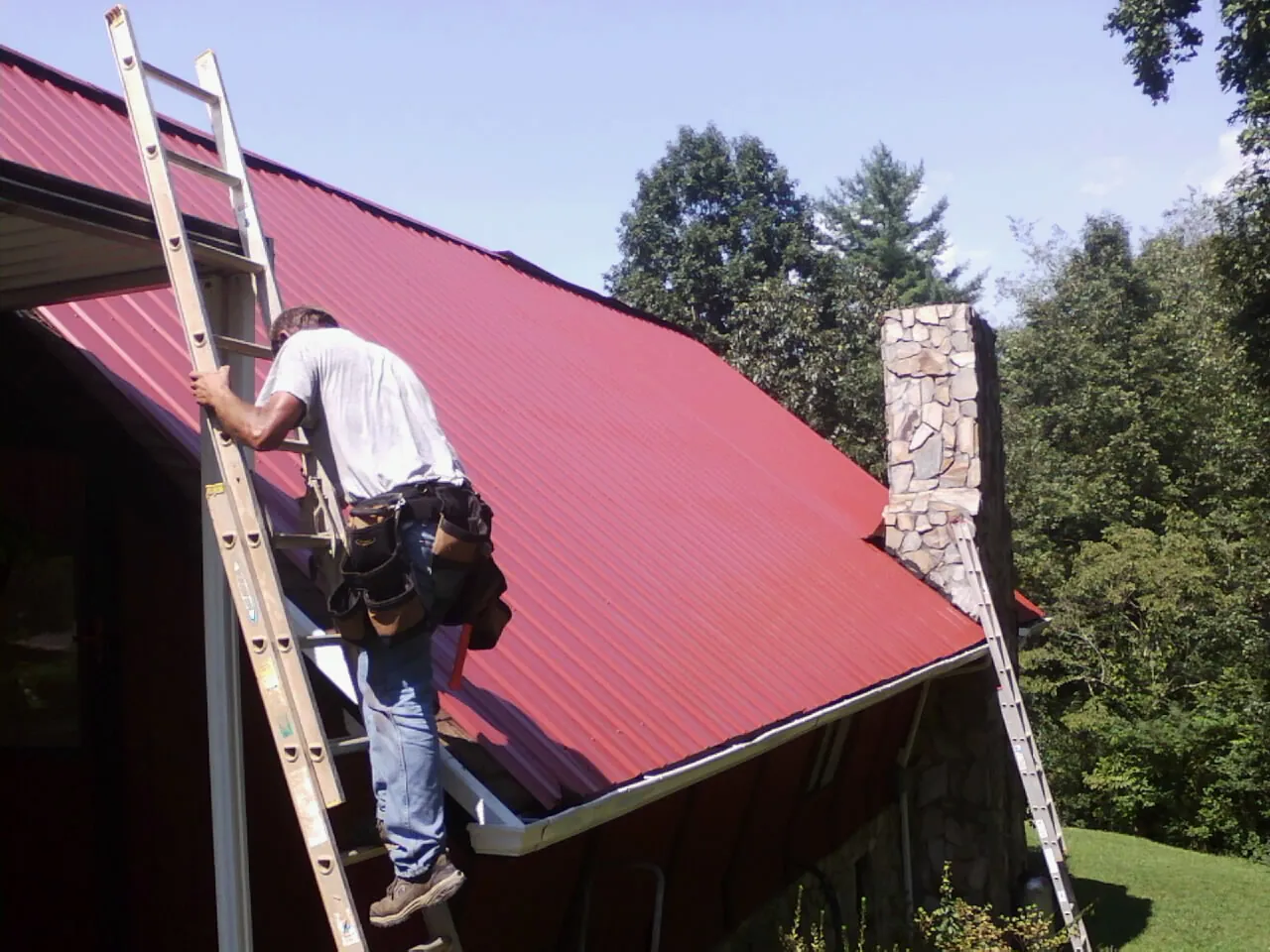 Roofer on a ladder beside a red metal roof and stone chimney — Soffit & Fascia Repair by Roofing Brothers in Mooreton